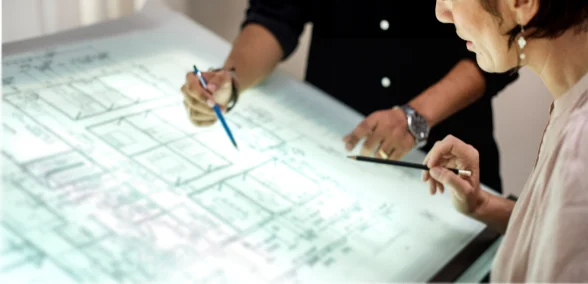 Zwei Personen prüfen gemeinsam einen technischen Gebäudeplan auf einem beleuchteten Tisch und markieren Details mit Stiften