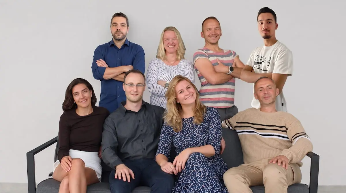 Teamfoto der SEO Revolution GmbH: Sieben lächelnde Teammitglieder, vier stehend im Hintergrund und vier sitzend auf einer grauen Bank vor einer hellen Wand.