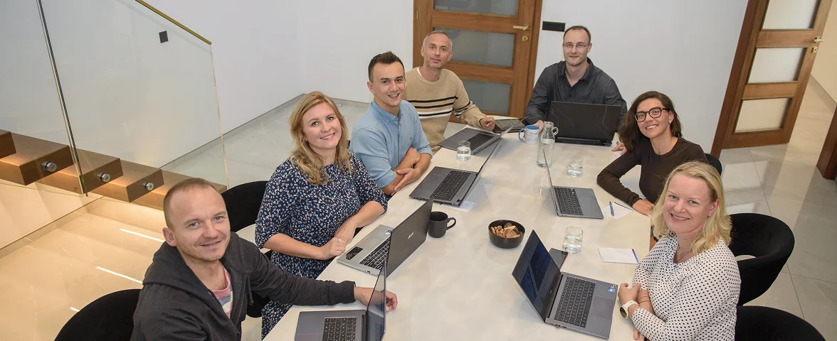 Team sitzt gemeinsam am Konferenztisch und schaut in die Kamera, freundliche Atmosphäre