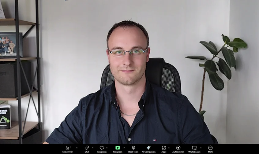 Mann sitzt im Büro vor dem Computer während eines Online-Meetings, freundlicher Blick in die Kamera