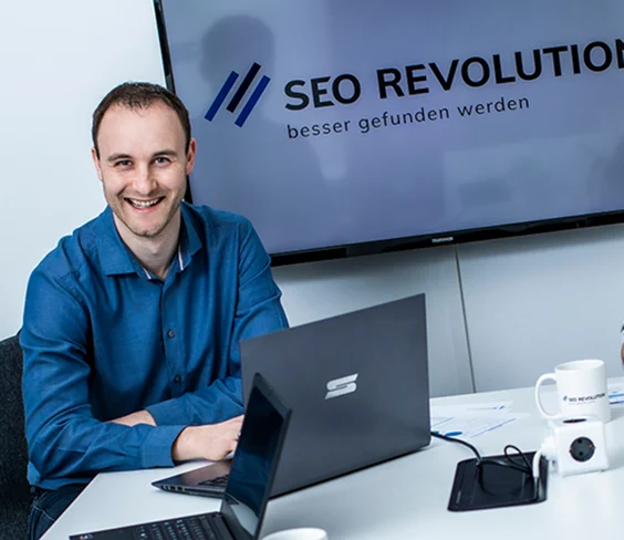 Mann sitzt lächelnd am Schreibtisch vor einem Monitor mit dem SEO Revolution Logo im Hintergrund