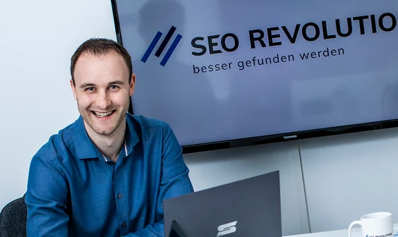Toni sitzt vor Laptop und Bildschirm mit SEO Revolution Logo und lächelt in die Kamera